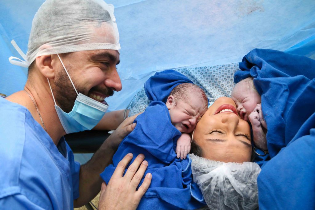 Fotografia de Parto em Curitiba: mãe sorridente deitada na maca com dois bebês recém-nascidos encostados em seu rosto, enquanto o pai, usando máscara e touca cirúrgica, sorri emocionado ao lado.