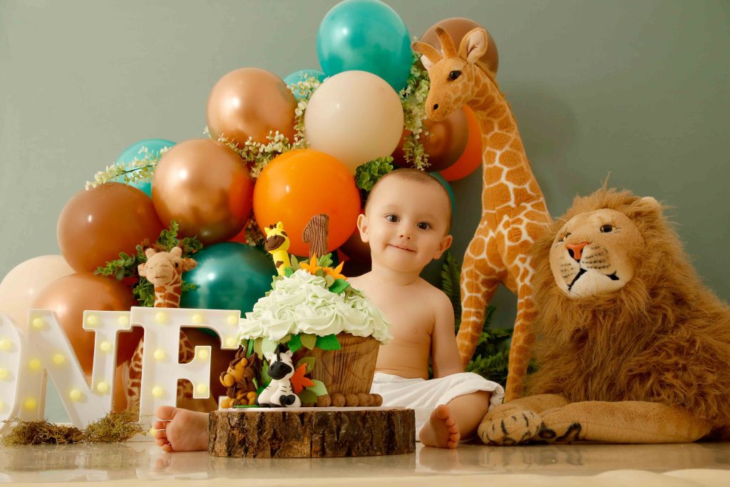 Bebê sorridente em ensaio Smash the Cake com tema safari, rodeado de bichinhos de pelúcia, bolo decorado e balões coloridos.