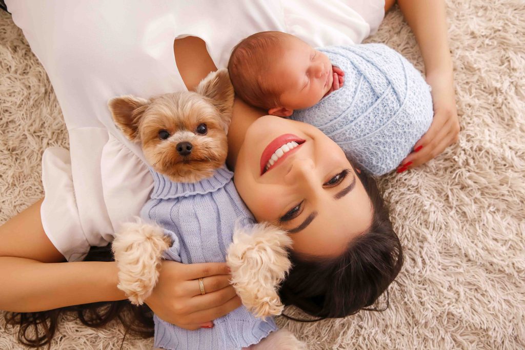 Mãe deitada no tapete abraçando bebê recém-nascido enrolado em manta azul e cachorrinho de roupa azul em ensaio newborn em Curitiba.